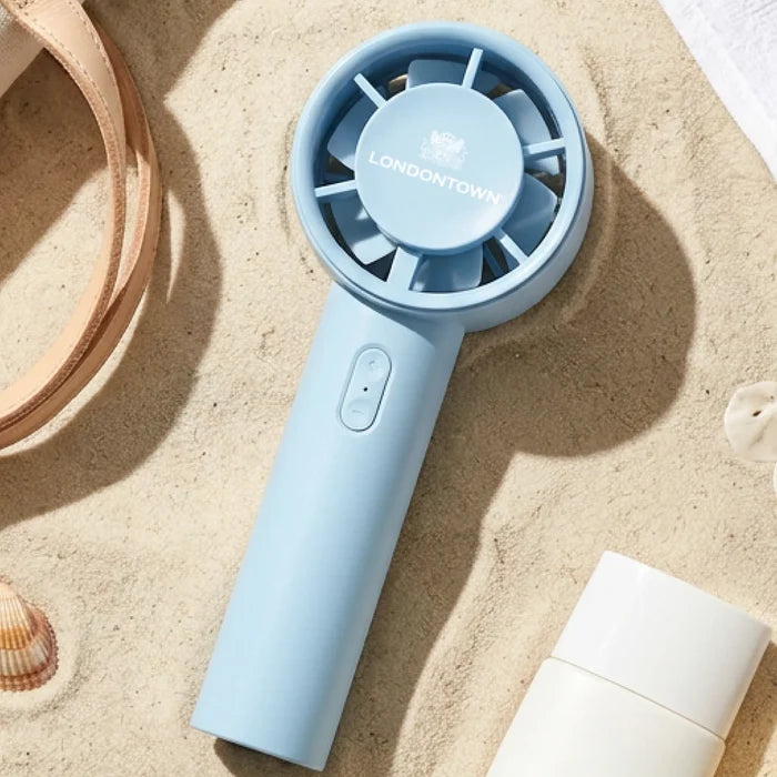 A light blue Cool Set Nail Fan - Ocean from LONDONTOWN rests on the sand near a bottle, a strap, seashells, and a towelβideal for beach days when you want to speed up nail dry time.