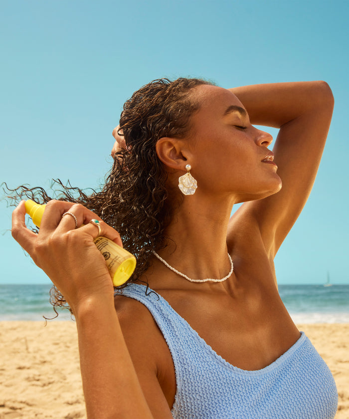 Woman with wet curly hair wearing a blue knitted tank top sprays a yellow bottle on her hair at a sunny beach.