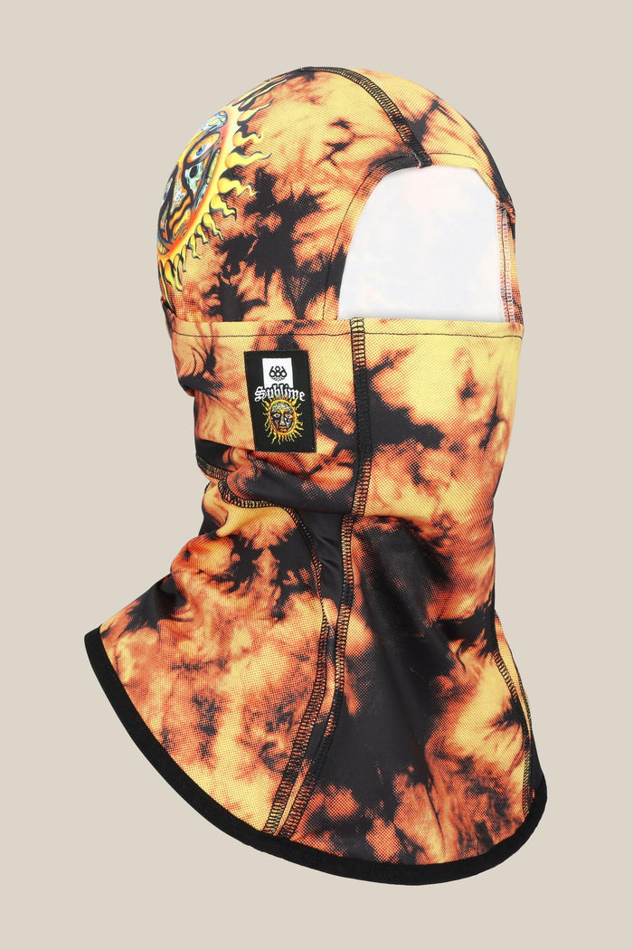 The 686 Alpha Hinged Balaclava by 686 features a black and orange tie-dye design, an eye within a sun graphic on the side, plus a small black patch with a sun logo and SENSITIVE CONTENT text.