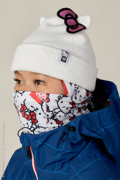 A person in a white knit hat with a Hello Kitty bow, a matching Hello Kitty neck gaiter, and a blue geometric jacket completes the look with the 686 Alpha Hinged Balaclava for extra warmth and style.