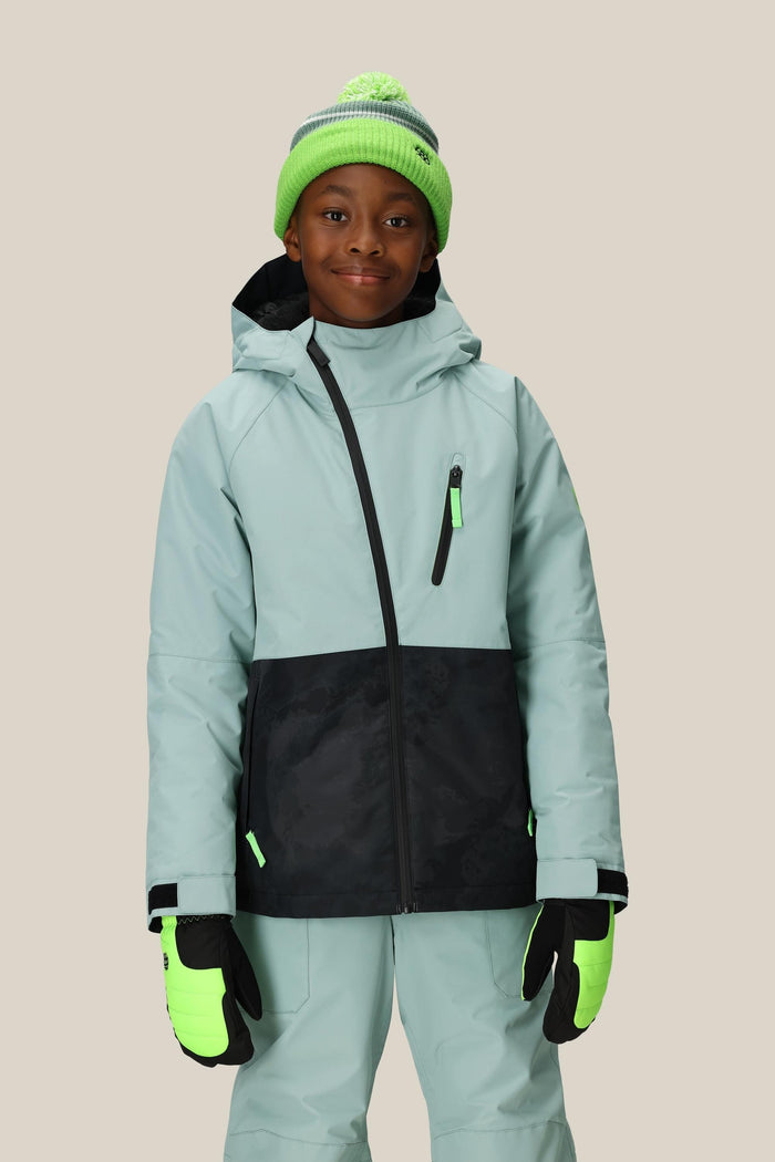 A child wearing the 686 Boys' Hydra™ Insulated Jacket by 686 in light blue with black accents, matching pants, neon green gloves, and a green pom-pom knit hat stands smiling against a beige background.