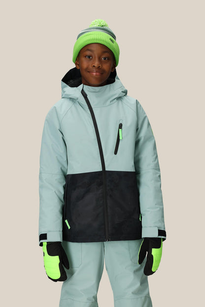 A child wearing the 686 Boys' Hydra™ Insulated Jacket by 686 in light blue with black accents, matching pants, neon green gloves, and a green pom-pom knit hat stands smiling against a beige background.