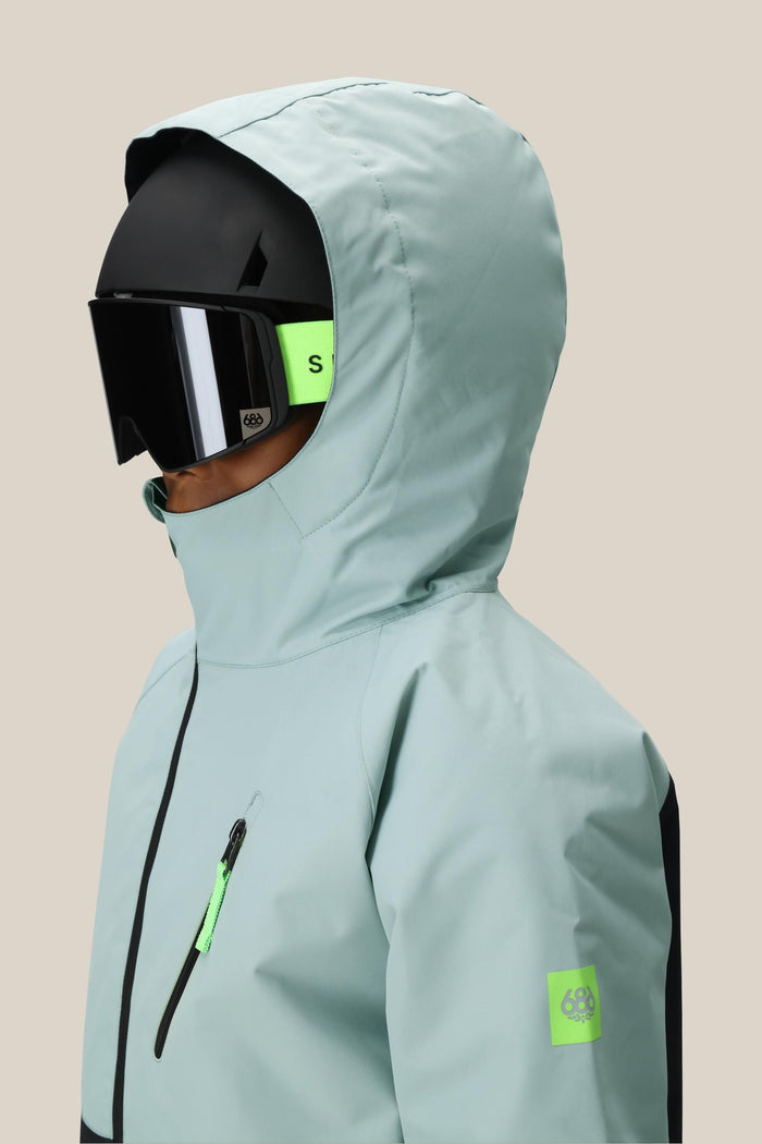 A boy wears the 686 Boys' Hydra™ Insulated Jacket in light blue with neon green accents, paired with a black ski helmet and large black ski goggles, shown in profile against a plain beige background. Brand: 686.