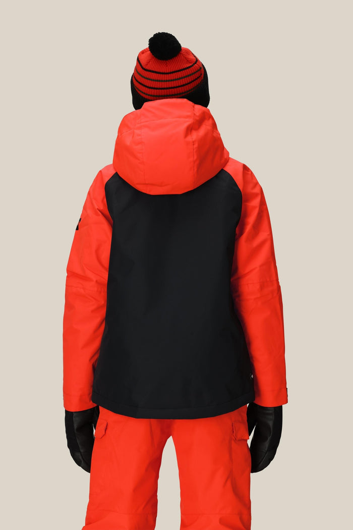 A person stands facing away, wearing the 686 Boys' Hydra™ Insulated Jacket by 686 in red and black with a hood, paired with red snow pants, black gloves, and a red and black striped pom-pom beanie against a beige background.