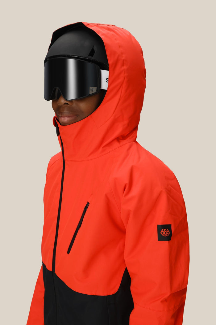 A person wears the 686 Boys' Hydra™ Insulated Jacket in bright red and black, paired with black ski goggles and a helmet, standing against a beige background. The jacket features a logo patch on the sleeve and a zippered chest pocket.