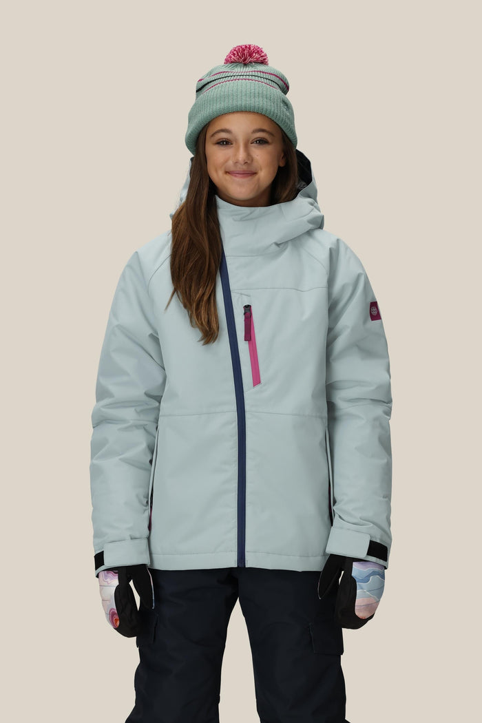 A girl smiles for the camera in the 686 Girls' Hydra™ Insulated Jacket by 686, paired with colorful gloves and a green beanie with a pink pompom, set against a plain beige background.
