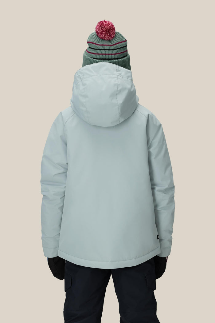 A person wearing the 686 Girls' Hydra™ Insulated Jacket by 686, paired with a green beanie with a red pom-pom, black gloves, and dark pants, stands with their back to the camera against a beige background.