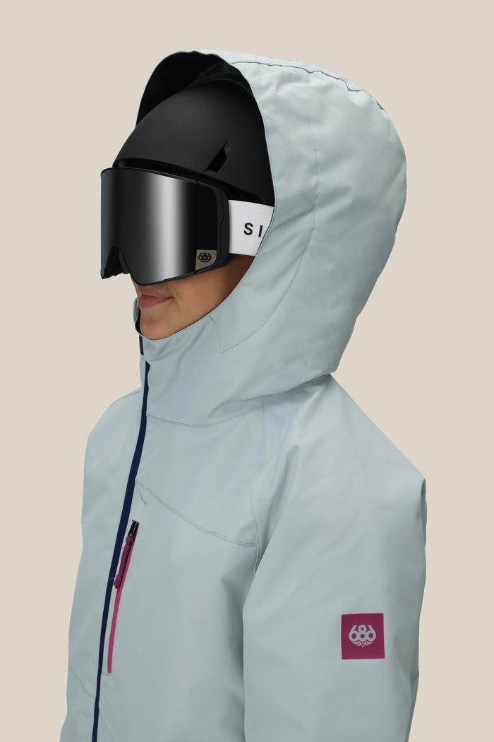 A girl wearing the 686 Girls' Hydra™ Insulated Jacket and black ski helmet with large black goggles, shown in profile against a plain beige background.