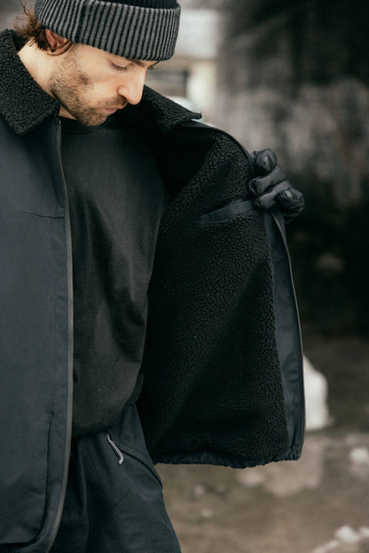 A person wearing the 686 Mens Caim Insulated Jacket holds it open outdoors to reveal its soft, furry lining. Theyre also wearing black gloves and a striped beanie, with a blurred outdoor background.