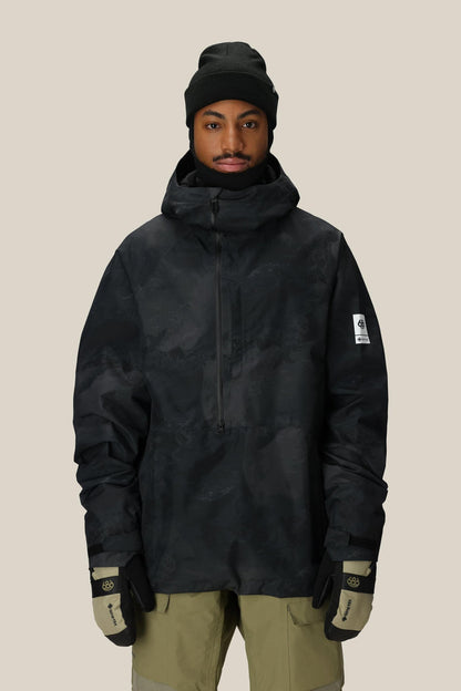 A person wearing the 686 Mens GORE-TEX Fragment Shell Anorak, black beanie, beige pants, and black-and-beige gloves stands against a plain light background.
