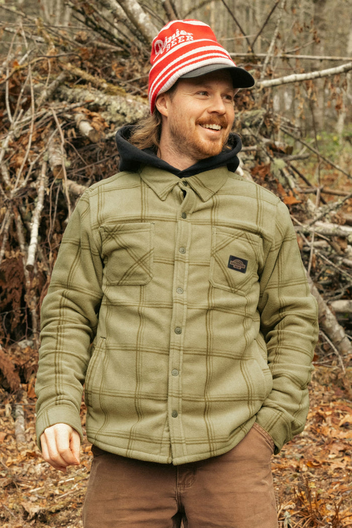 A man smiles outdoors among branches and dry leaves, wearing the 686 Men's Sierra Fleece Flannel by 686, paired with a red and white striped beanie, olive green quilted jacket, black hoodie, and brown pants.