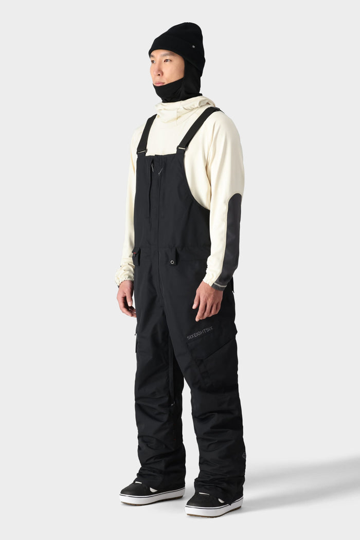 A person wears a cream hoodie, black beanie, black neck gaiter, and 686 Men's SMARTY® 3-in-1 Cargo Bib by 686 with removable liner, standing slightly right against a plain light gray background.