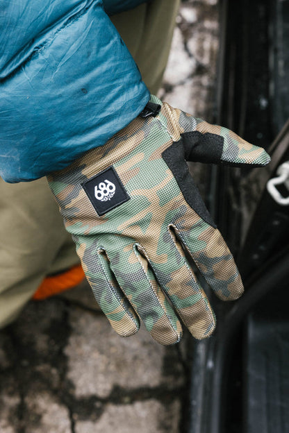 A person wearing a blue jacket and 686 Outlaw Pipe Glove by 686 stands next to a car on cracked pavement.