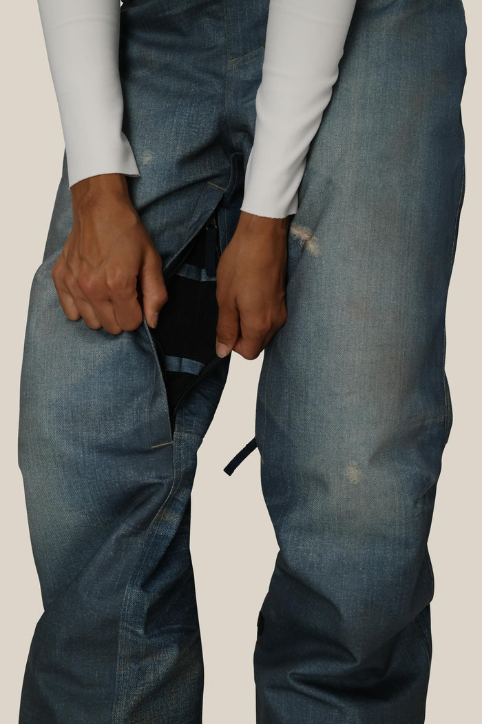A person wearing a white long-sleeve shirt and blue jeans unzips the front of their pants, revealing 686 Women's Deconstructed™ Denim Insulated Pants by 686, made from durable waterproof polyester fabric underneath.