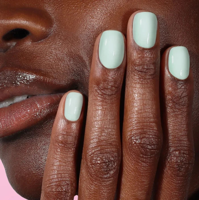 A close-up of a person’s face and hand with glossy lips and nails painted in LONDONTOWN’s Beary Minty shade, set against a pink background.