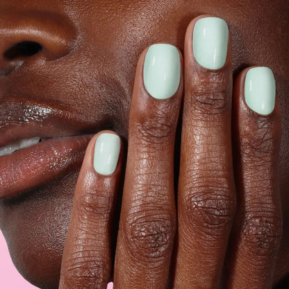 A close-up of a person’s face and hand with glossy lips and nails painted in LONDONTOWN’s Beary Minty shade, set against a pink background.