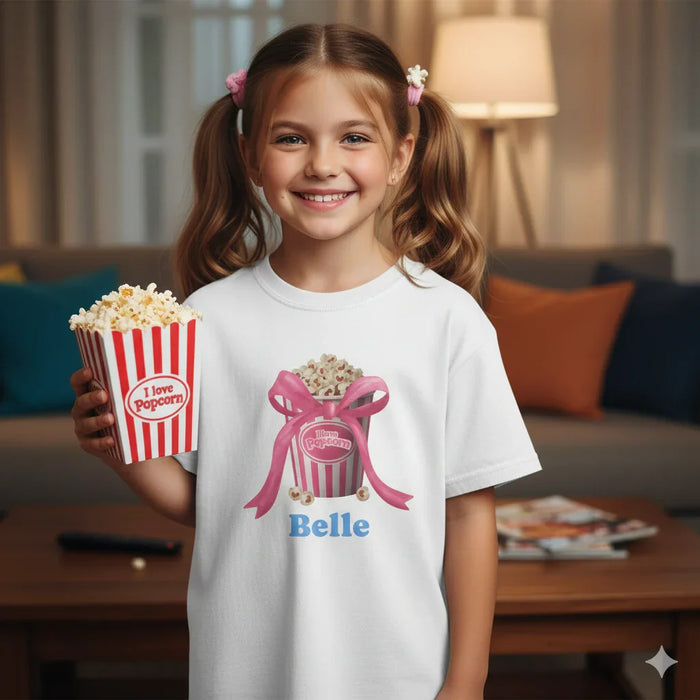Smiling young girl with pigtails holding popcorn in red-white striped box, wearing white popcorn-themed T-shirt with name Belle