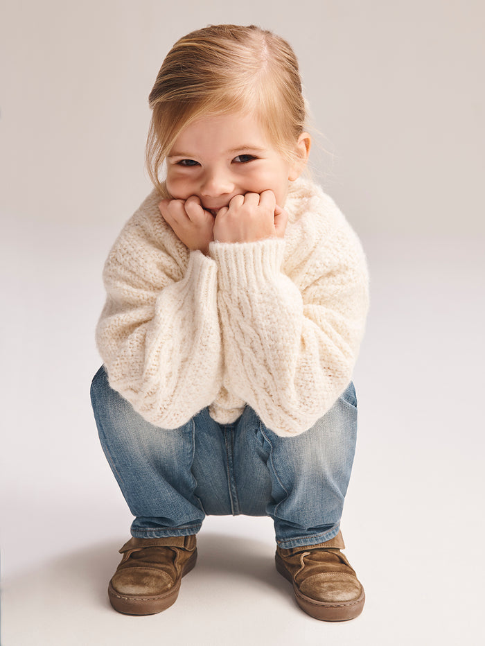 DL1961 boys' Brady Slim jeans in light blue 'Fresh', worn by a smiling child crouching in a cream sweater and tan boots.