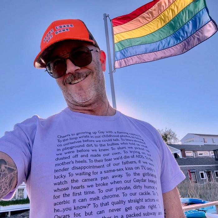 Adam of Adam's Nest wearing lilac Leo Herrera Cheers to Growing Up Gay t-shirt on Provincetown beach with rainbow flag,
