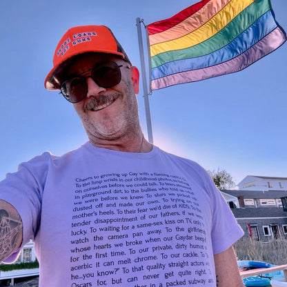 Adam of Adam's Nest wearing lilac Leo Herrera Cheers to Growing Up Gay t-shirt on Provincetown beach with rainbow flag,