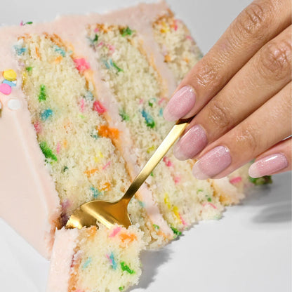A hand with pastel pink nails uses a gold fork to cut into a slice of LONDONTOWN Confetti Cake, featuring white frosting and colorful sprinkles.