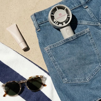 The LONDONTOWN Cool Set Nail Fan - Cloud peeks out from a back jeans pocket, alongside a tube of cream, sunglasses, and a striped towel scattered on the sandy surface—perfect essentials for those dry times at the beach.
