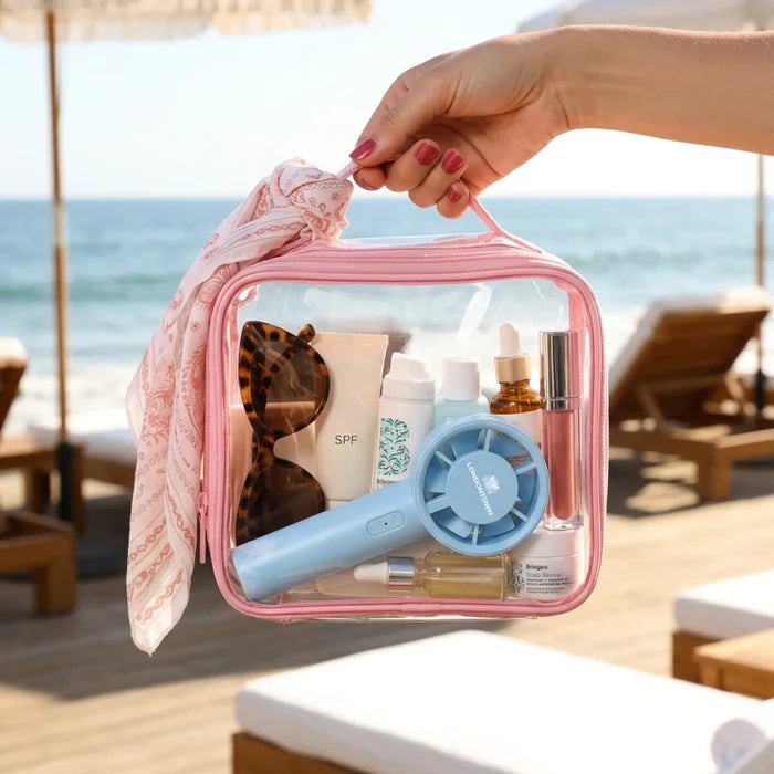 A hand holds a clear pink bag containing sunglasses, sunscreen, skincare products, and the LONDONTOWN Cool Set Nail Fan in Ocean on a beach with lounge chairs and umbrellas in the background.