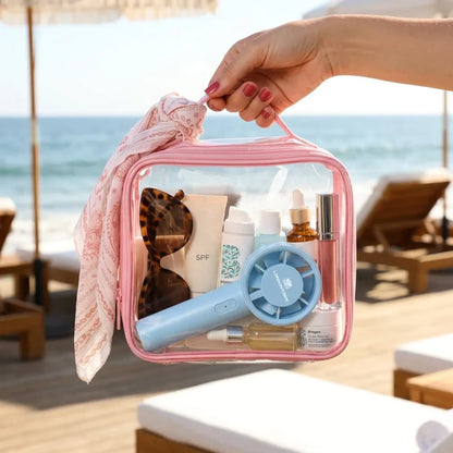 A hand holds a clear pink bag containing sunglasses, sunscreen, skincare products, and the LONDONTOWN Cool Set Nail Fan in Ocean on a beach with lounge chairs and umbrellas in the background.