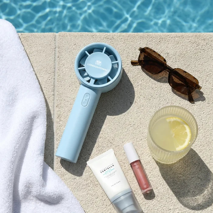 Poolside scene featuring the LONDONTOWN Cool Set Nail Fan - Ocean alongside sunglasses, a lemon-infused drink, a white towel, and three cosmetic items on a stone surface next to the swimming pool—perfect for achieving shorter nail polish dry times under the sun.