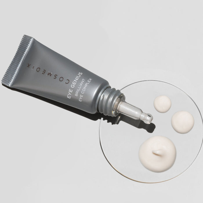 Cosmedix Eye Genius Brilliant Eye Complex tube with dripping light beige serum on glass petri dish against light gray background