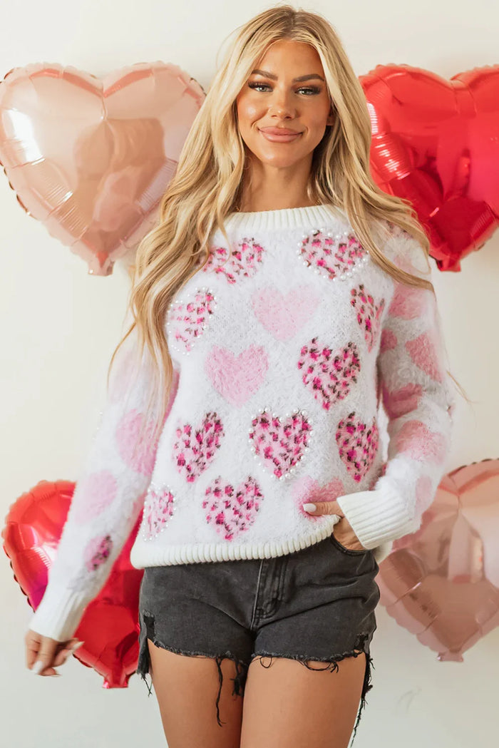Woman wearing a white sweater with pink heart patterns, standing against a light background with heart-shaped balloons.