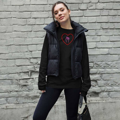 Woman wearing a black puffer vest over a black shirt with a heart and pink solidarity fist design, standing against a brick wall.