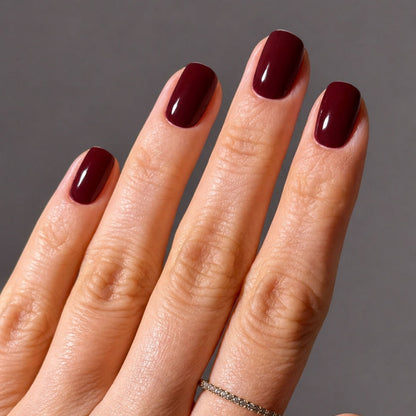 A hand with short, neatly manicured nails painted in LONDONTOWN’s Red Velvet, a decadent deep burgundy polish, wearing a thin silver ring on the ring finger, set against a plain gray background.