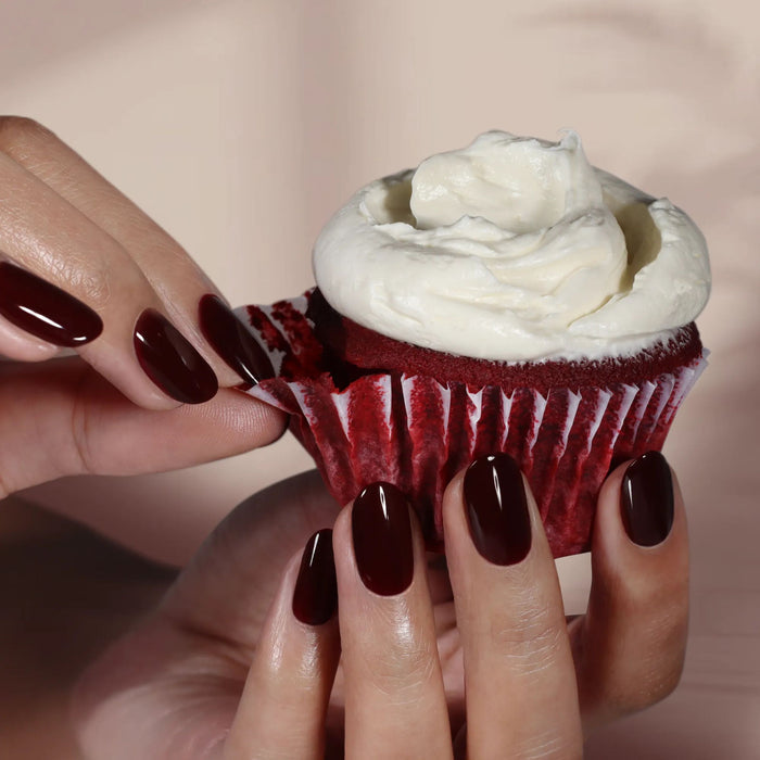 Hands with LONDONTOWN’s Red Velvet polish—a deep burgundy shade—hold a decadent red velvet cupcake topped with white frosting, peeling back the cupcake liner.
