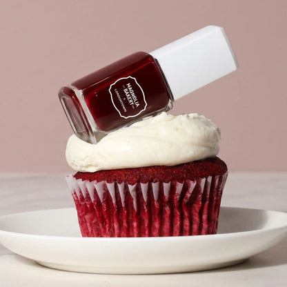 A bottle of LONDONTOWN’s Red Velvet nail polish rests on the frosting of a red velvet cupcake on a white plate, set against a plain pink background.