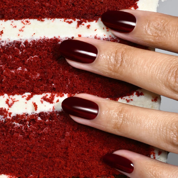A hand with decadent, deep burgundy nails painted in LONDONTOWN’s Red Velvet shade rests on a slice of red velvet cake with white frosting layers.