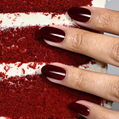 A hand with decadent, deep burgundy nails painted in LONDONTOWN’s Red Velvet shade rests on a slice of red velvet cake with white frosting layers.