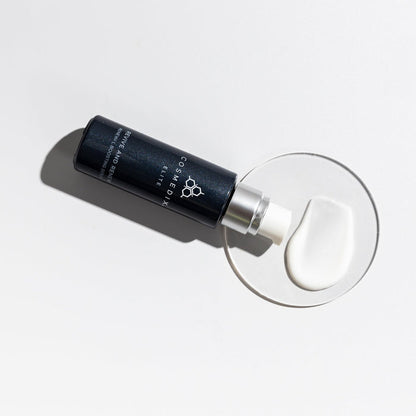 CosMedix Elite Renewal Boosting Serum bottle with dropper laid on a white surface beside glassware