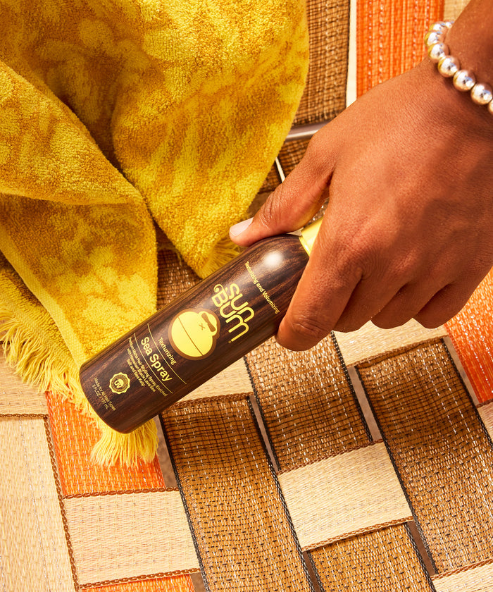 A hand with a pearl bracelet holding a dark brown bottle of sea spray hair product over a woven textured mat with orange, brown, and cream colors, and a yellow textured towel nearby.