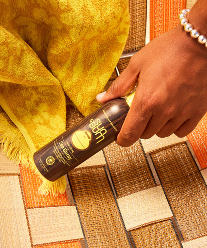 A hand with a pearl bracelet holding a dark brown bottle of sea spray hair product over a woven textured mat with orange, brown, and cream colors, and a yellow textured towel nearby.