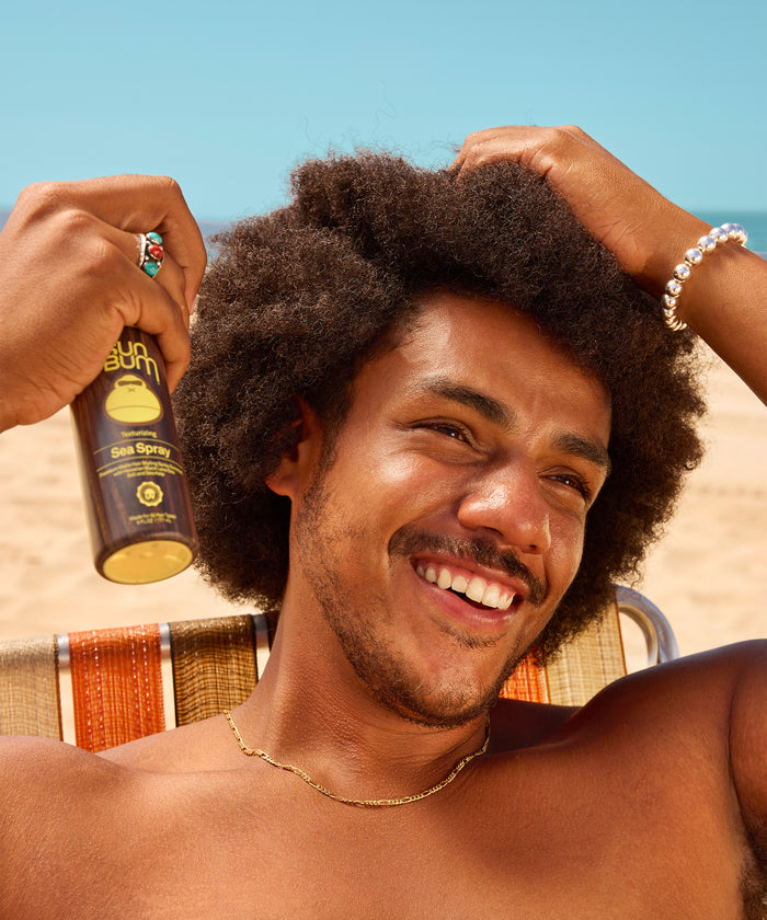 Smiling man with curly hair applies sea spray hair product while sitting outdoors on a sunny day with a clear blue sky.