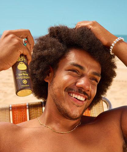 Smiling man with curly hair applies sea spray hair product while sitting outdoors on a sunny day with a clear blue sky.