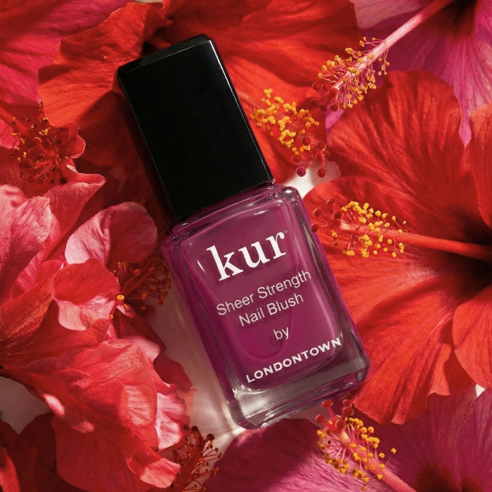 A bottle of LONDONTOWN Sheer Strength Nail Blush - Hibiscus, a nourishing nail hardener, sits among vibrant red hibiscus flowers.