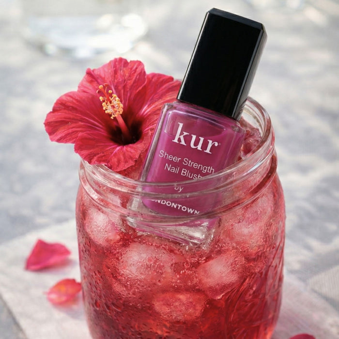 A bottle of LONDONTOWN Sheer Strength Nail Blush - Hibiscus sits in a mason jar filled with ice and red liquid, topped with a red hibiscus flower.
