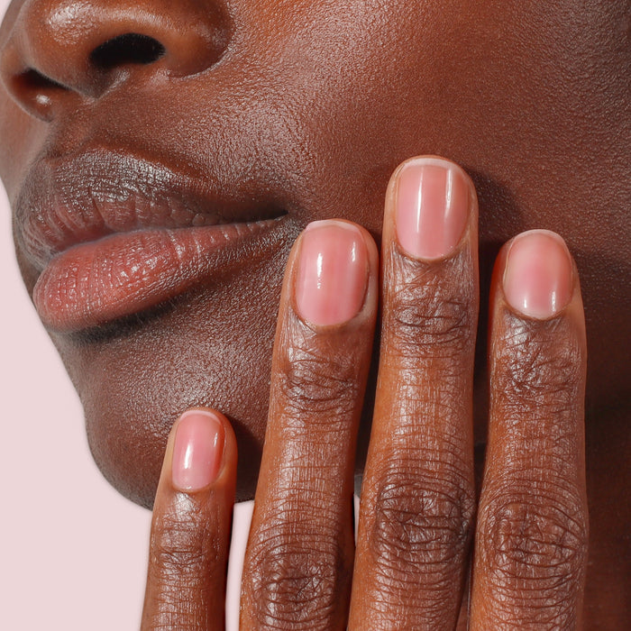 Close-up of a person's face and hand with smooth, glowing skin and neatly manicured nails enhanced by LONDONTOWN Sheer Strength Nail Blush in Hibiscus, set against a light pink background.
