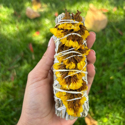 Hand holding a smudge stick with yellow flowers and white string against a grassy background